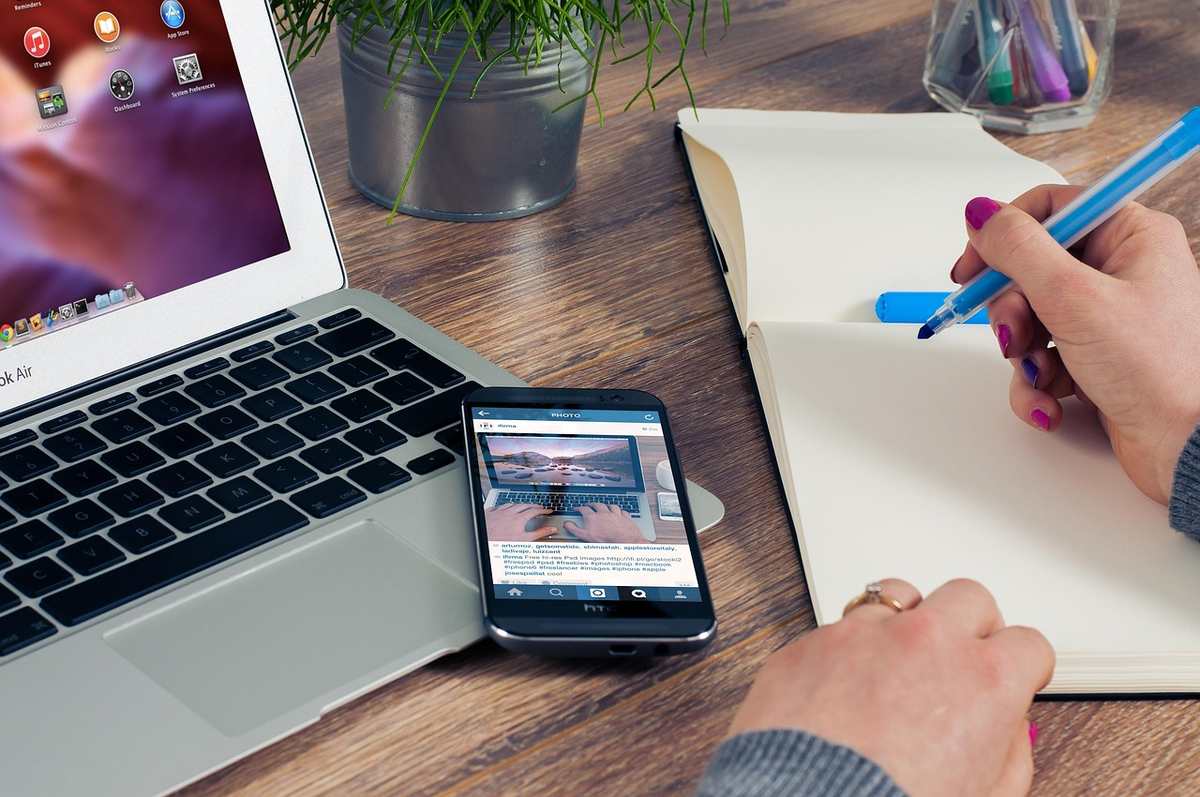 Une main écrit dans un cahier à côté d'un ordinateur portable et d'un smartphone Une main écrit dans un cahier à côté d'un ordinateur portable et d'un smartphone